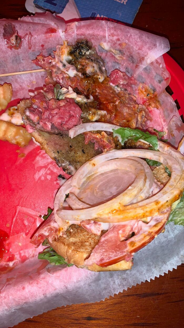 Unappetizing food picture showing a messy burger with raw meat, wilted lettuce, onions, and sauces on a red tray.