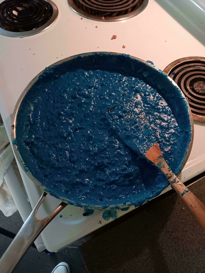 Pan filled with unappetizing blue thick food mixture on stove with wooden spatula stuck inside the pan.