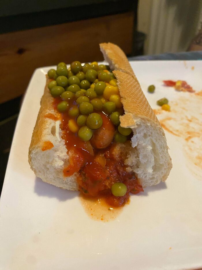 Unholy food pic of a hot dog topped with peas and sauce on a white plate, looking unappetizing and messy.