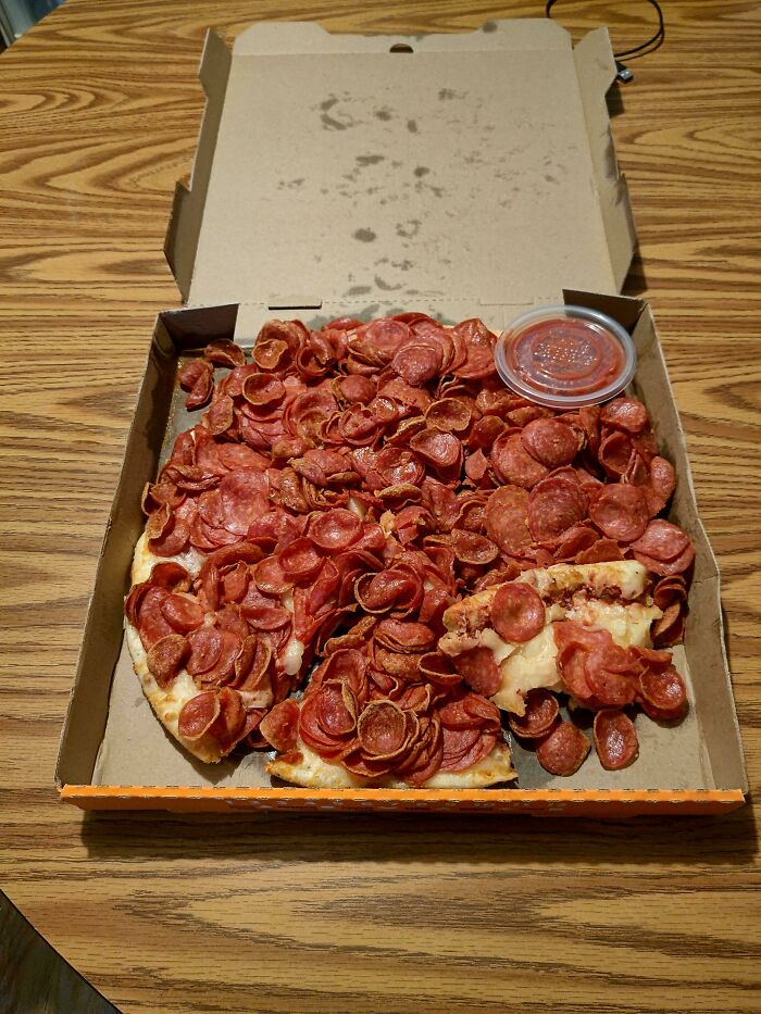 Pizza overloaded with crispy, curled pepperoni slices in a greasy box with a small container of marinara sauce.