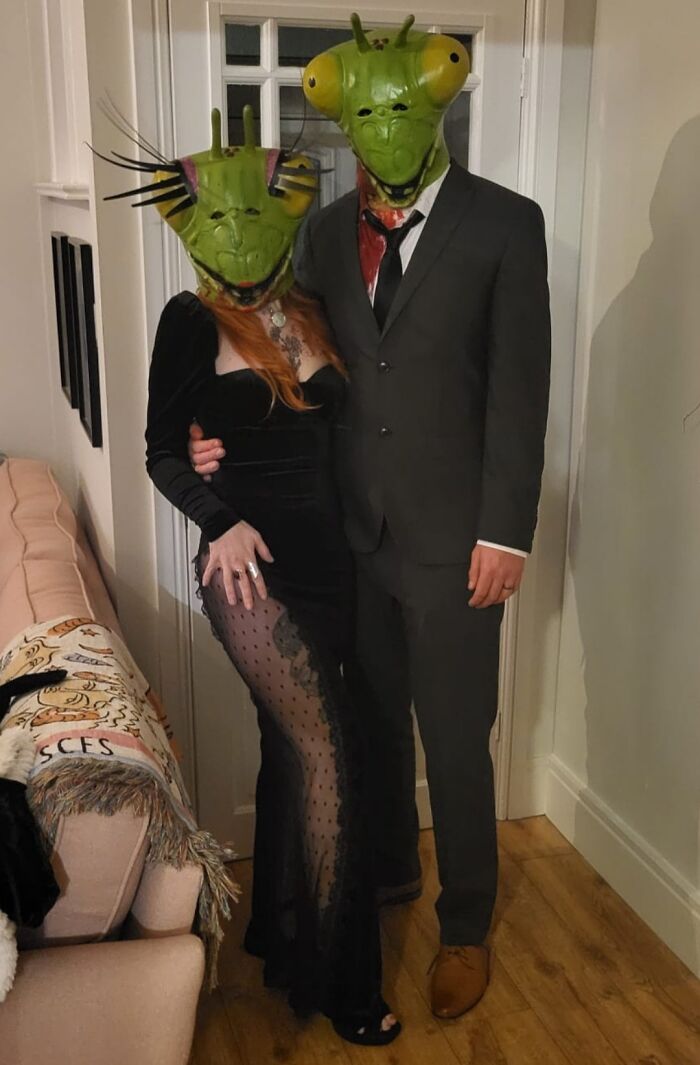 Couple in genius Halloween costumes wearing green insect masks with formal attire indoors at a party setting.