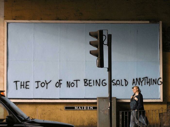 Graffiti on a billboard reading the joy of not being sold anything with a pedestrian walking past in an urban setting about simple living.