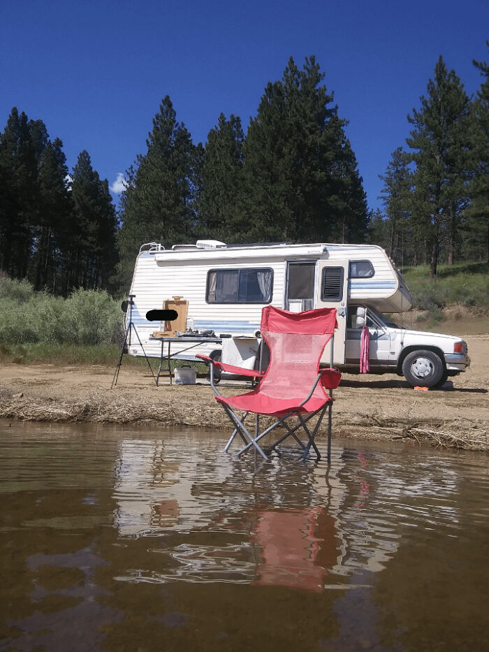 Red camping chair by lakeside with a camper van and trees in the background, illustrating simple living outdoors.