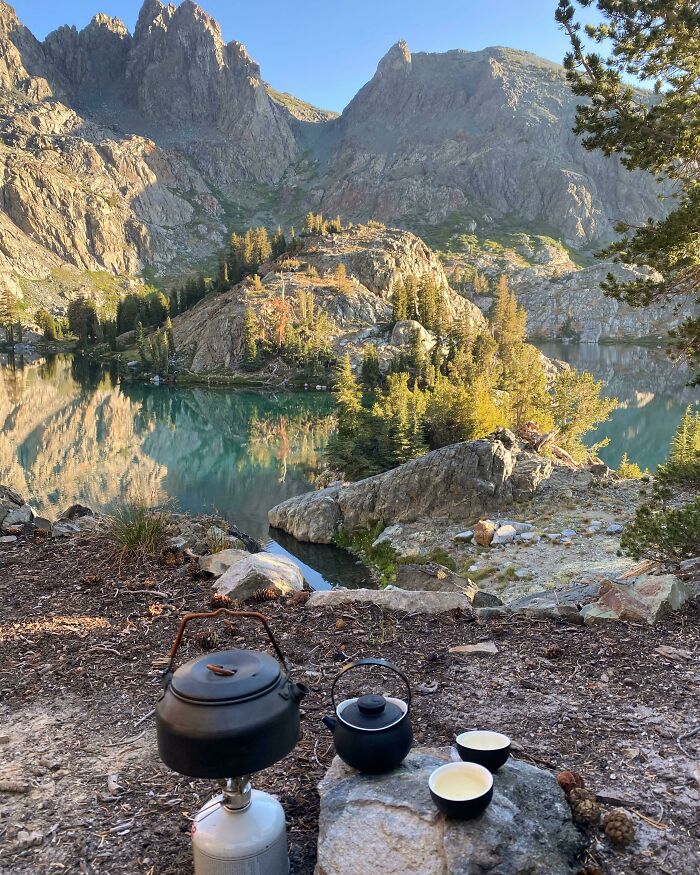 Camping tea setup with kettle and cups overlooking a mountain lake surrounded by rocky peaks and trees in simple living style.