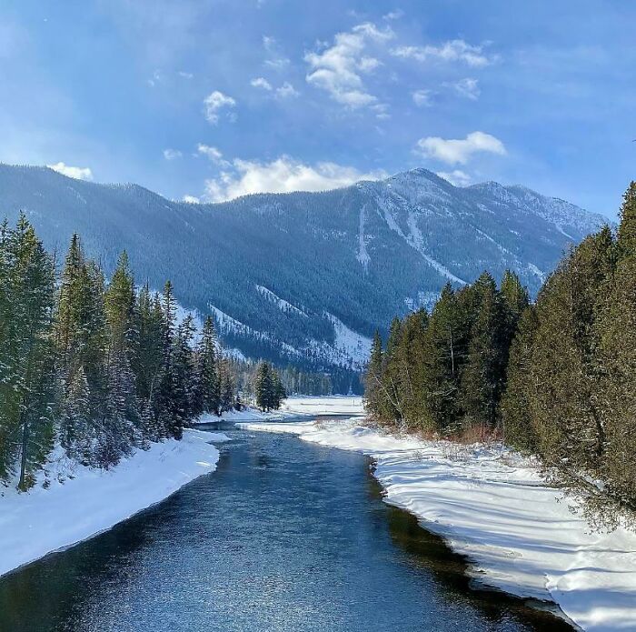 Snow-covered river landscape with evergreen trees and mountains under a blue sky, reflecting simple living and nature’s calm.