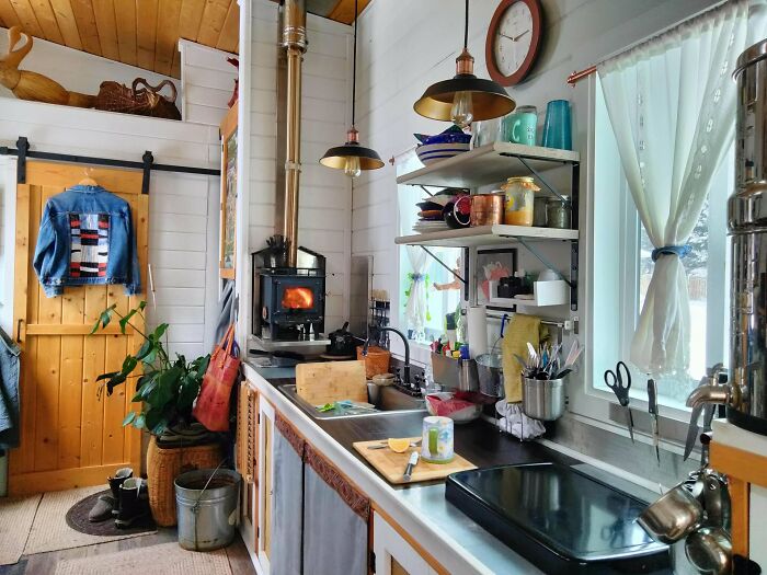 Cozy simple living kitchen with wood stove, open shelving, natural light, and minimalistic decor in a small home setting.