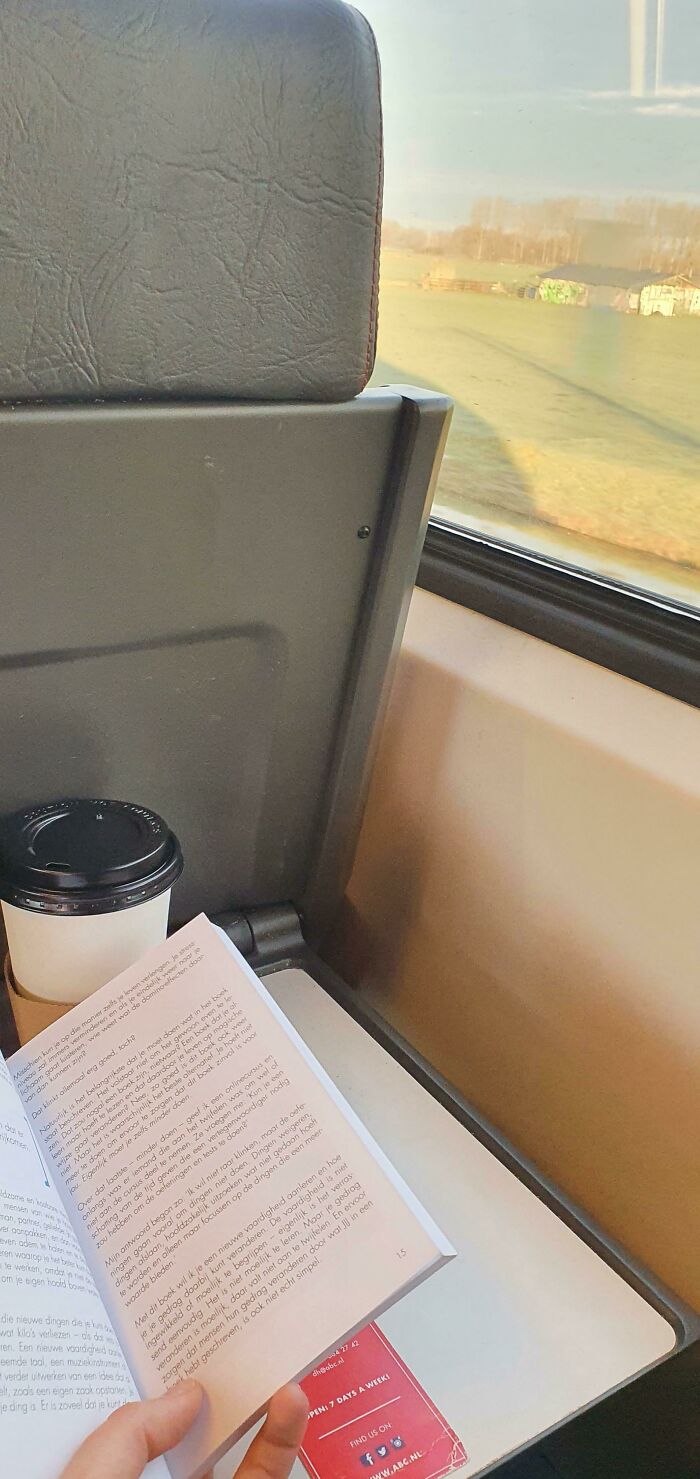 Hand holding book and coffee cup on train seatback tray, enjoying simple living and mindful moments on a journey.