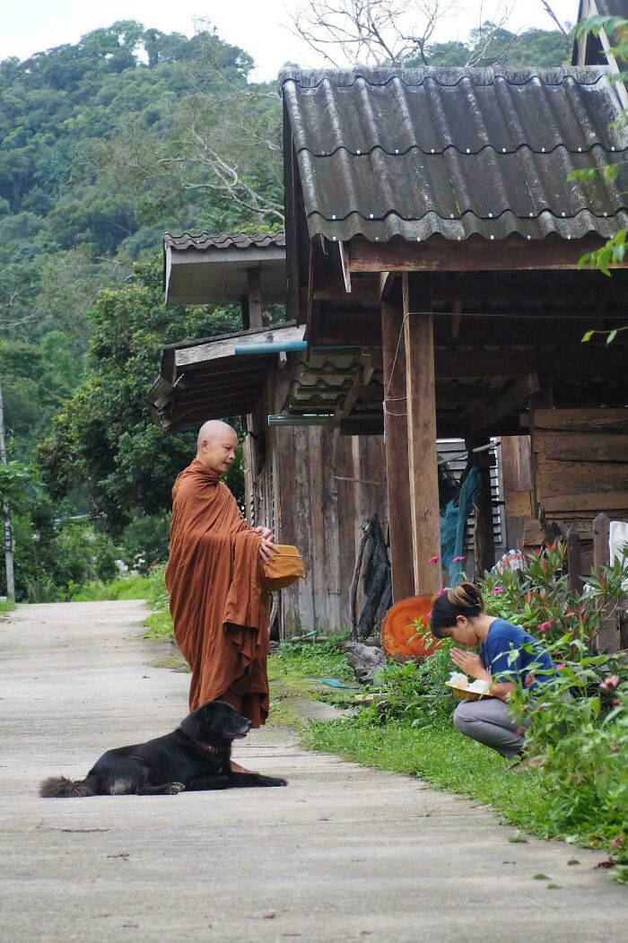 Monk receiving alms from a woman outside a rustic wooden house in a peaceful simple living setting.
