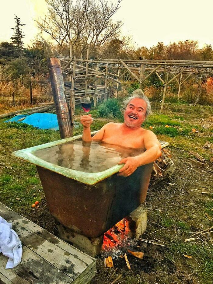 Man enjoying simple living in an outdoor rustic bathtub heated by fire, holding a glass of wine and smiling contentedly.