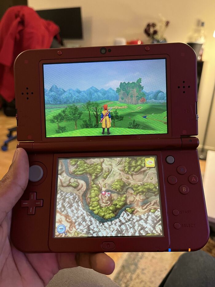 Hand holding a red Nintendo 3DS showing a game with a character in a simple living environment and a detailed map below.