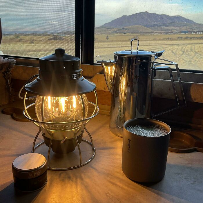 Cozy simple living scene with a lit lantern, metal coffee pot, and cup by a window overlooking a mountain landscape.