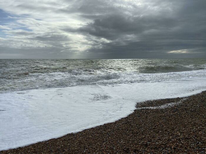 Calm ocean waves gently washing over a pebble beach under a cloudy sky, evoking simple living and peaceful moments.