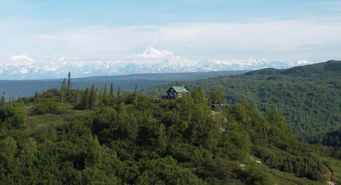 Remote cabin nestled among trees on a green mountain with distant snowy peaks under a clear blue sky, symbolizing simple living.