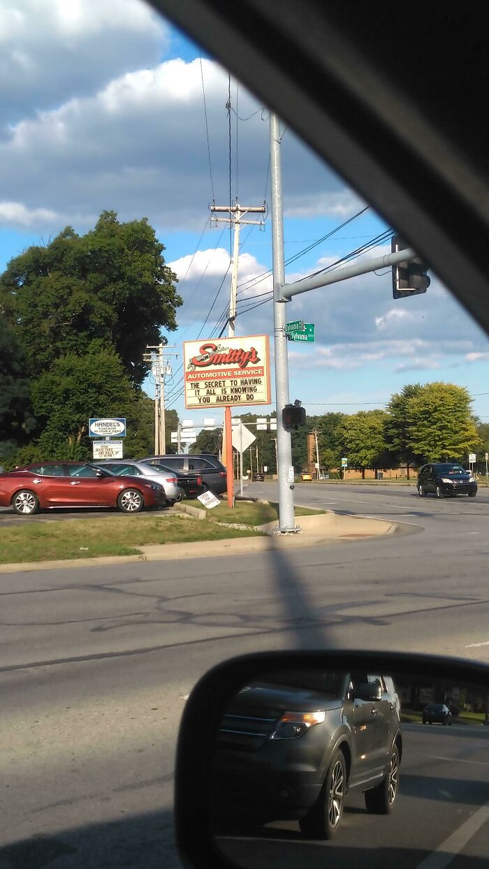 Street view with cars, trees, and a sign for automotive service, captured from inside a vehicle, highlighting simple living outdoors.