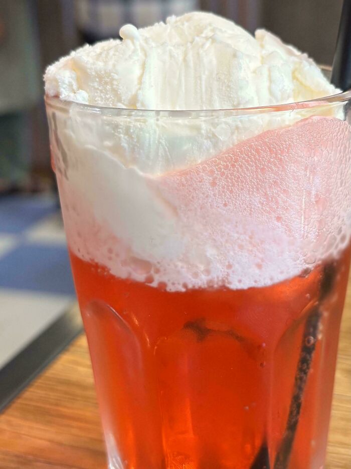 Glass of red fizzy drink topped with whipped cream resting on a wooden table, capturing simple living moments.