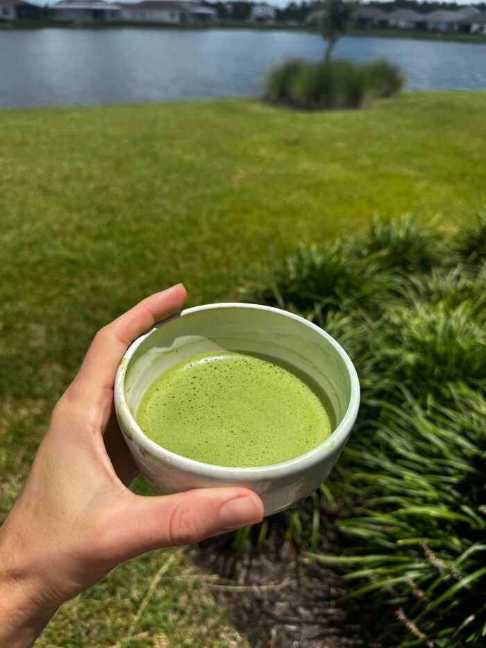 Hand holding a bowl of green matcha tea outdoors near water and garden, reflecting simple living and minimalism lifestyle.