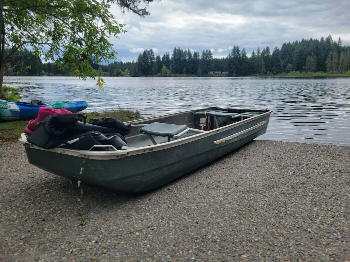 A simple living scene with a small boat on the shore beside calm water and a forested background under cloudy skies.