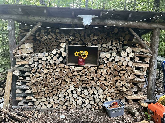 Neatly stacked firewood under a rustic shelter with sunflowers in a jar, illustrating simple living and minimalism.