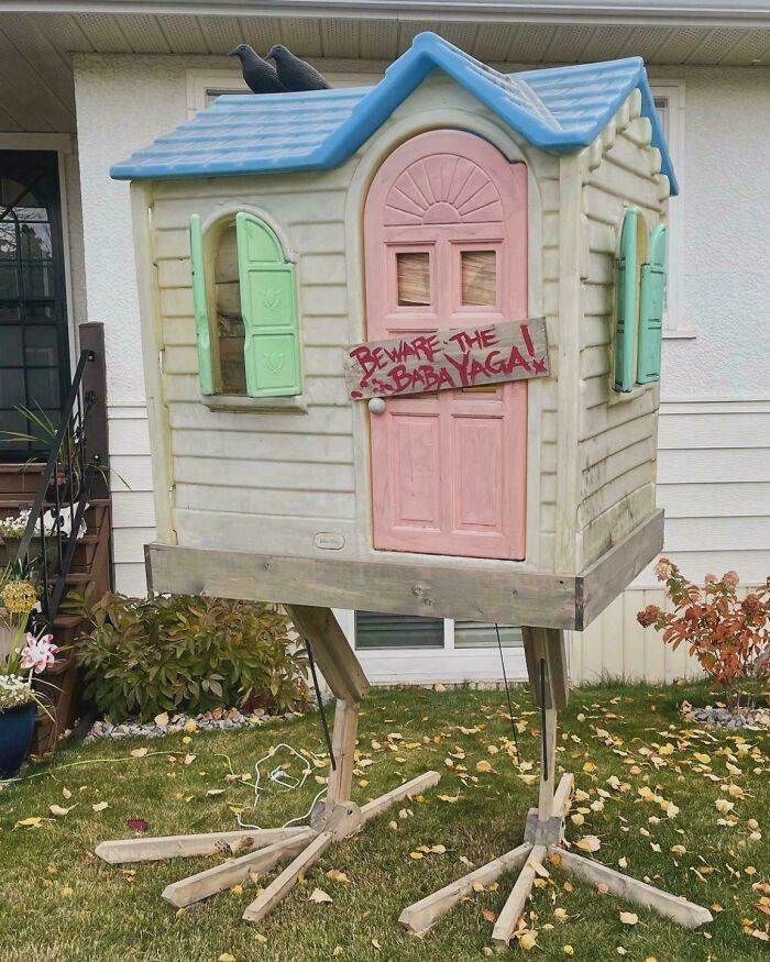 Outdoor Halloween decoration of a small elevated house with a Beware the Baba Yaga sign and crow props on the roof.