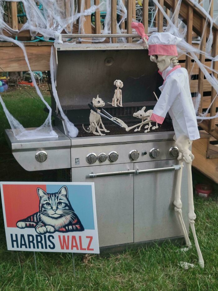 Skeleton dressed as a chef grilling small skeleton dogs on a decorated outdoor Halloween setup with cobwebs.