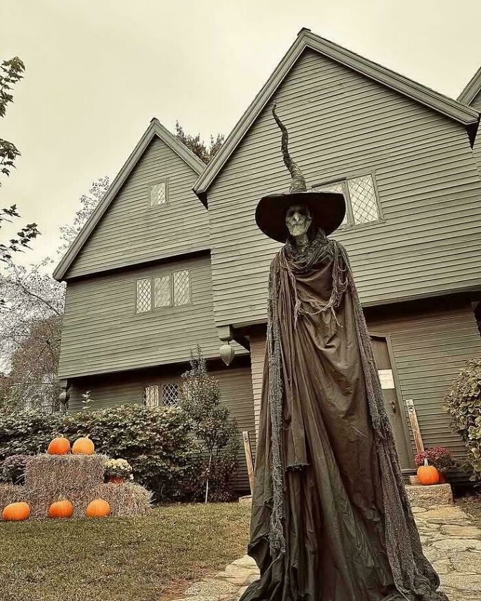 Tall witch skeleton outdoor Halloween decoration in front of a house with pumpkins and hay bales in yard.