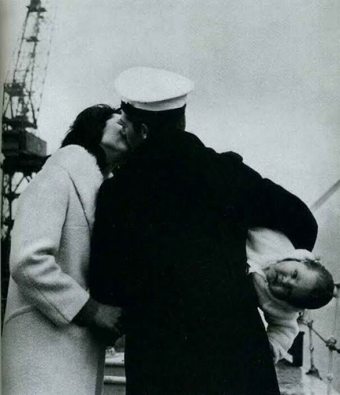Black and white photo of a sailor kissing a woman while holding a child, a fascinating moment to see the world differently.