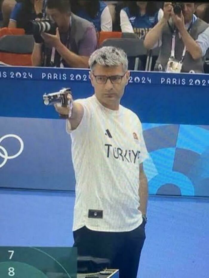 Man wearing a Turkey shirt aiming a pistol in a sports shooting event with photographers in the background.