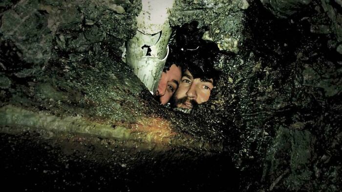Two men peering through a narrow opening in dark rocky terrain, featured in fascinating photos and stories.