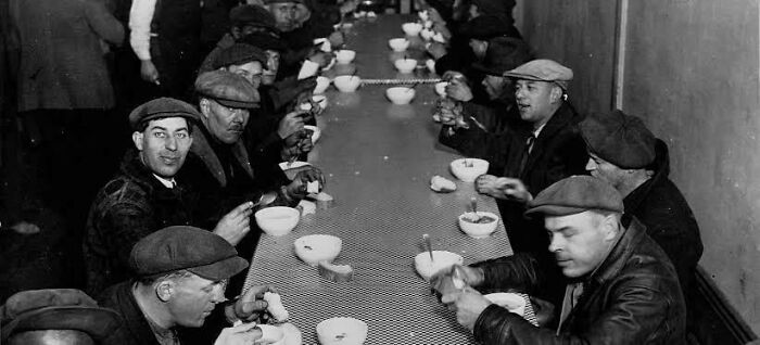 Black and white photo of men in caps sitting at a long table eating, capturing fascinating stories that may change how you see the world.