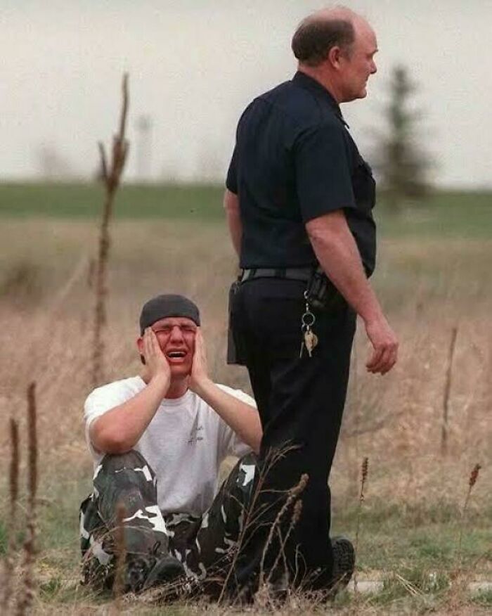 Young man crying on ground with hands on face while police officer stands nearby in field, a fascinating photo capturing raw emotion.