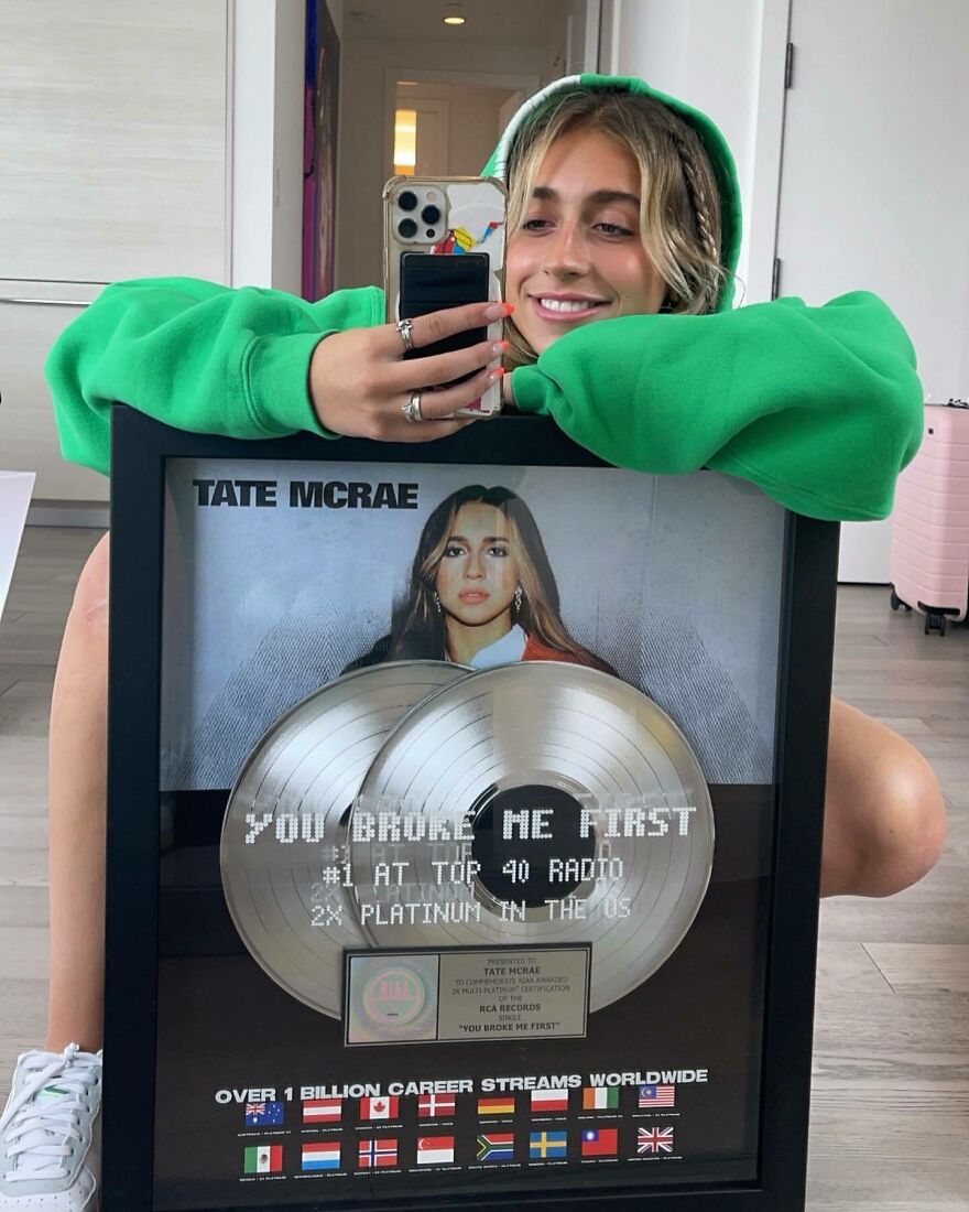 Tate Mcrae in a green hoodie holding a platinum record award for You Broke Me First, showcasing her music success. Tate Mcrae in a green hoodie holding a platinum record award for You Broke Me First, showcasing her music success.