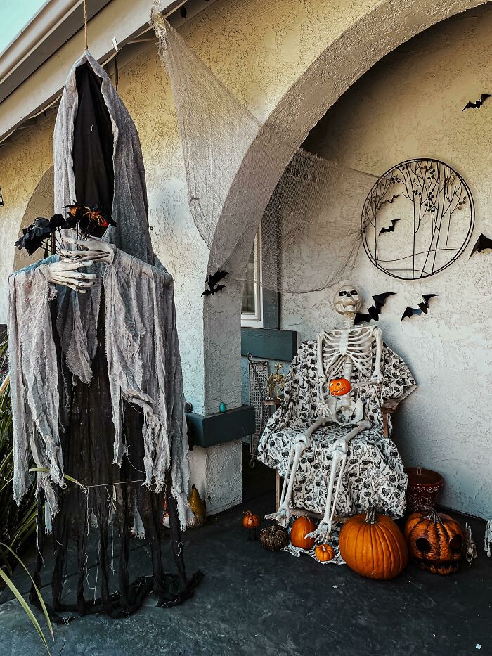 Outdoor Halloween decorations featuring spooky skeleton, pumpkins, hanging ghost figure, and black bat wall accents on porch.