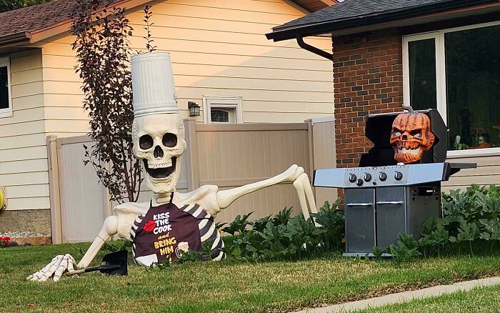 Large skeleton wearing a chef hat and apron by a barbecue grill with a carved pumpkin, outdoor Halloween decorations.