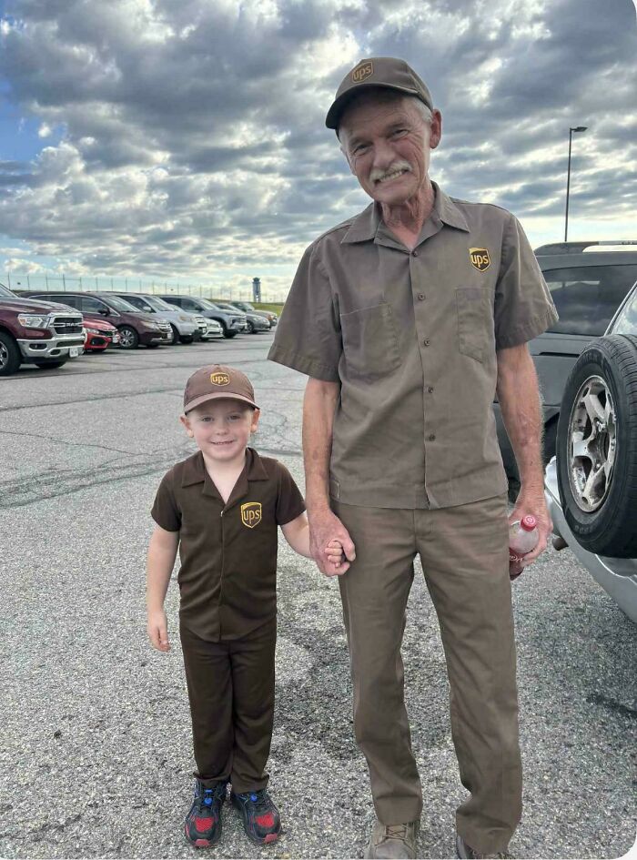 Older and young men in UPS uniforms holding hands, smiling together in a parking lot under a cloudy sky.