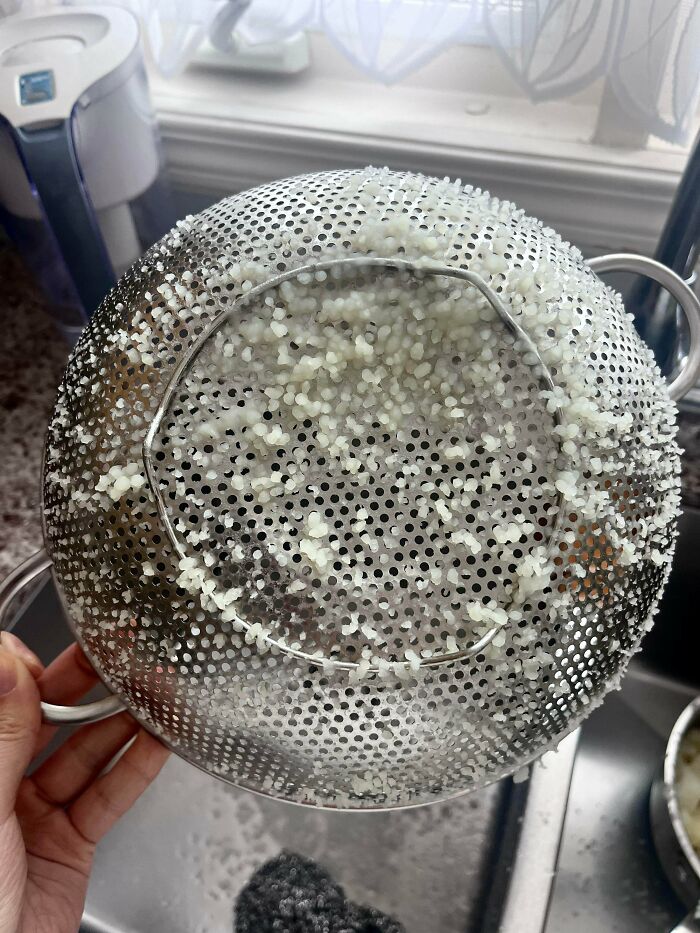 Metal colander with stuck rice grains creating a trypophobia-triggering pattern held over a kitchen sink.