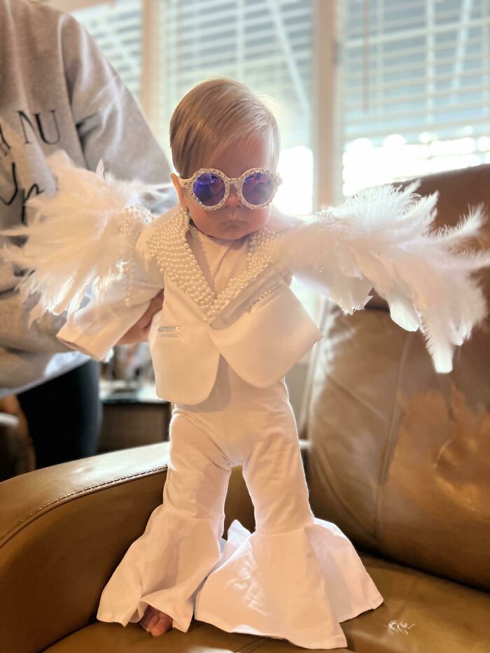 Toddler in an amazing kids' Halloween costume wearing white feathered wings, pearl jacket, and round sunglasses indoors.