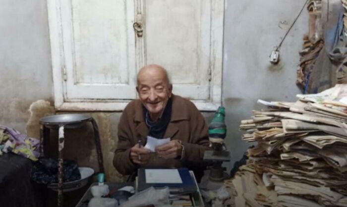 Elderly man smiling warmly while sorting papers in a cluttered room, showing the good in the world.