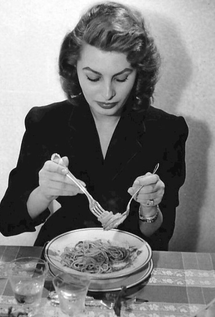 Sophia Loren, Italian beauty, twirling spaghetti with fork and spoon at a table, captured in a classic black and white photo.