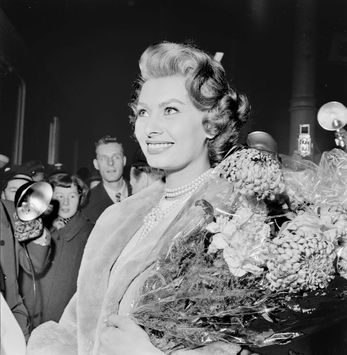 Sophia Loren smiling warmly, wearing pearls and holding flowers, surrounded by fans and photographers, showcasing Italian beauty.