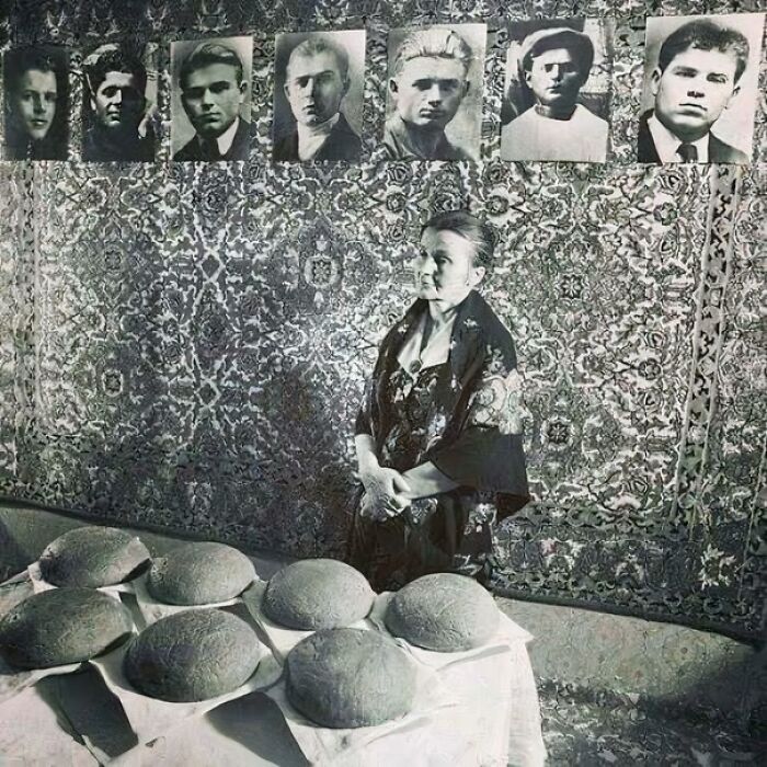 Woman standing beside a table with round bread loaves and black-and-white portraits on a patterned wall in a fascinating photo.