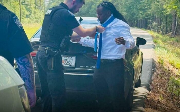 Police officer helping man by tying a necktie beside a car on a rural road, showing men are good deeds.