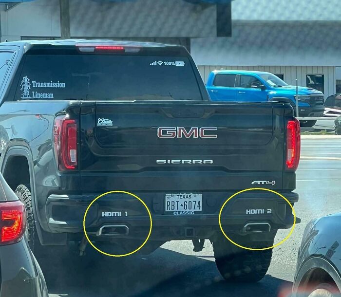 Black GMC Sierra pickup with humorous bumper stickers labeled HDMI 1 and HDMI 2 on the exhaust pipes, snapped on the road.