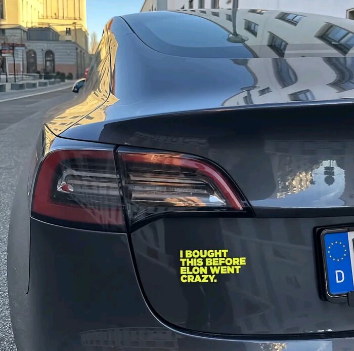 Black car with a clever and hilarious bumper sticker about Elon Musk spotted on the road in an urban setting.