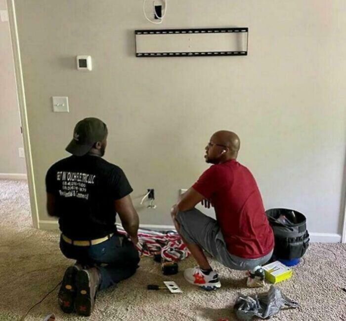 Two men kneeling on carpet, installing a TV mount bracket, demonstrating men are good through teamwork and kindness.