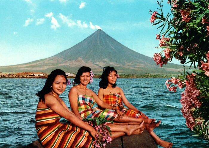 Three women in colorful dresses sit by the water near a volcano, capturing vintage vacation pics before cell phone cameras.