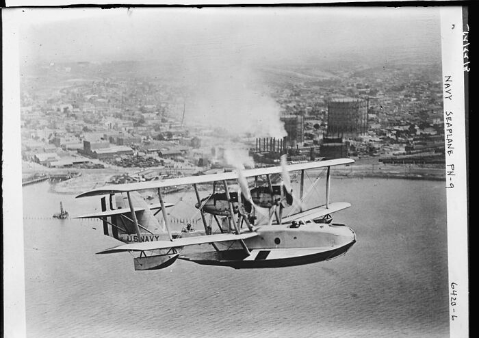 Vintage black and white photograph of a Navy seaplane flying over a coastal city, illustrating historical aviation stories.
