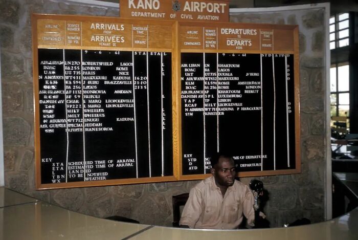 Vintage airport arrivals and departures board at Kano Airport with a man at the desk, showcasing vacation pics from before cell phone cameras.