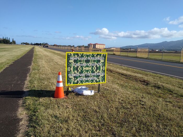Confusing road sign with camo pattern warns of heavy haul in progress, blending into the grassy roadside causing unclear instructions.