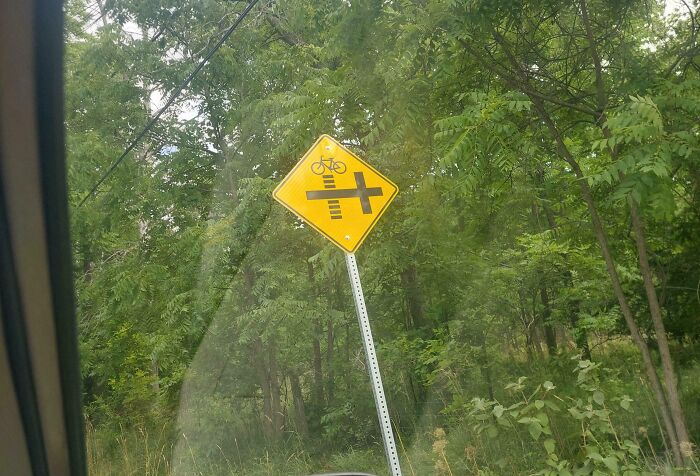 Confusing road sign warning of a bike crossing with unclear directions, captured from a car window surrounded by trees.