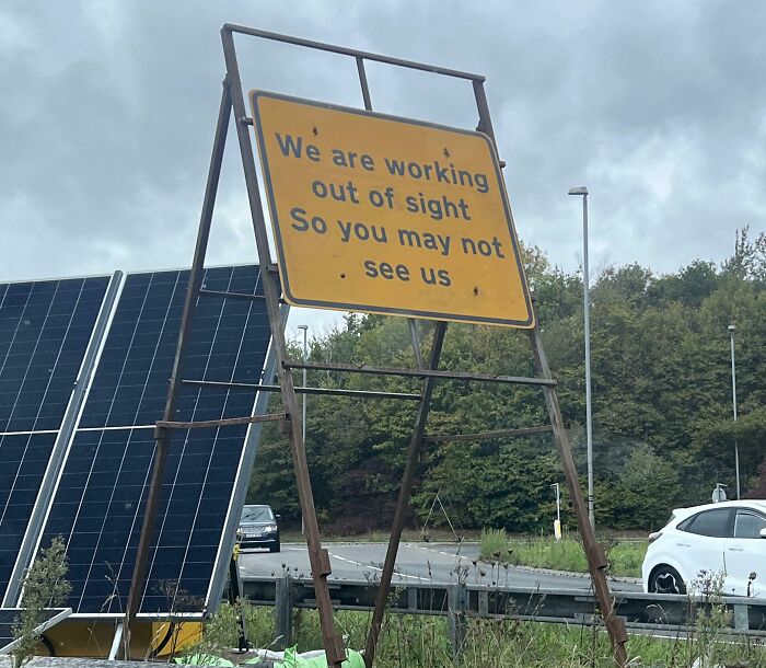 Yellow road sign on rusted frame reads we are working out of sight so you may not see us near highway with cars and solar panels